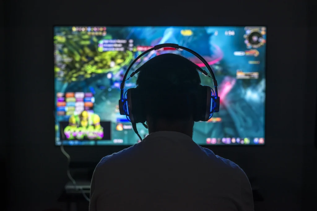 teen boy wearing headphone plays a video game in the dark, photo taken from rear creating a silhouette 