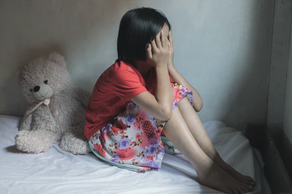 A sad and frightened girl sits alone in a dark room, her hands covering her face. She feels lonely, stressed, and scared.