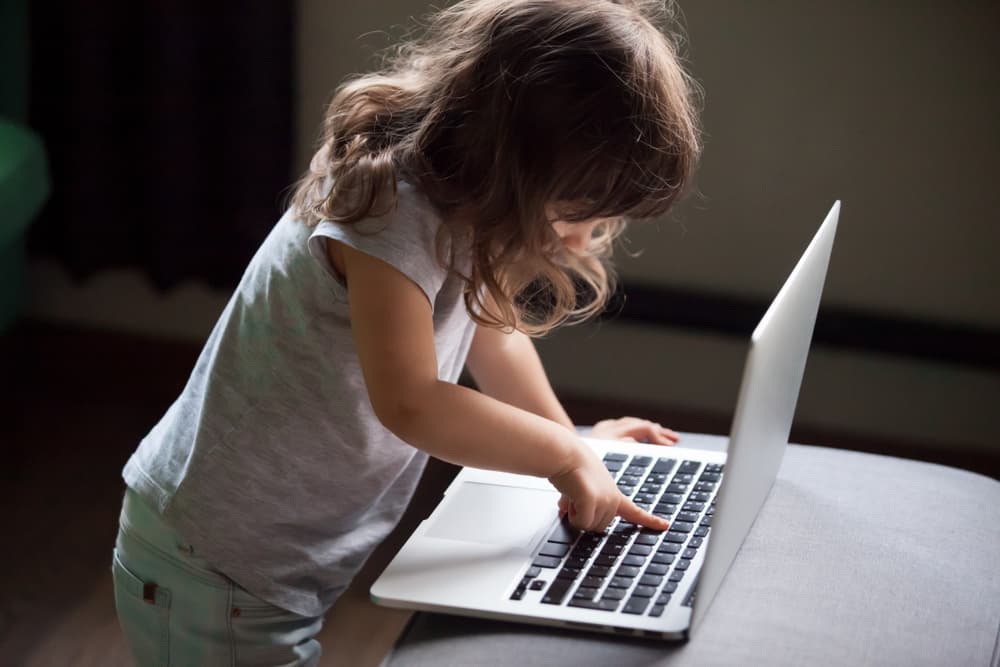 A curious and smart little girl is typing on a laptop alone. This clever child is using the computer online without permission, accessing forbidden internet content despite parental protections.