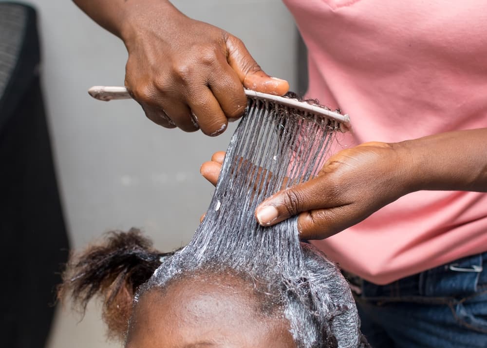 The hands of an African stylist apply relaxer cream with a comb to the natural, long hair of a female customer at a beauty salon.