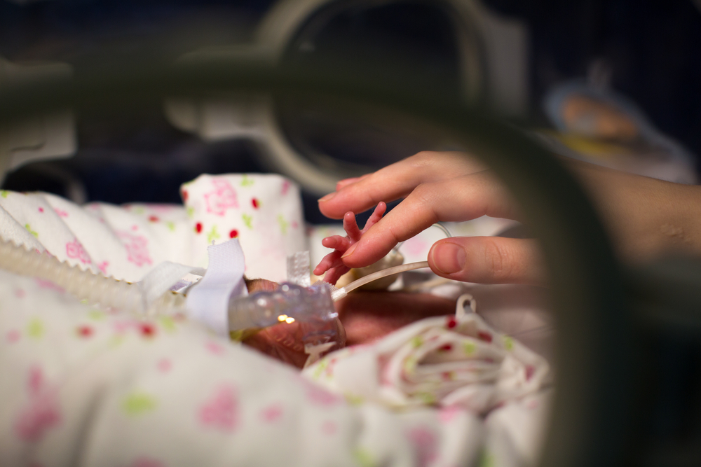 A close-up of a parent's hand touching a hospitalized baby's hand in an incubator, depicting the severe medical complications and rickets symptoms cited in Neocate litigation.